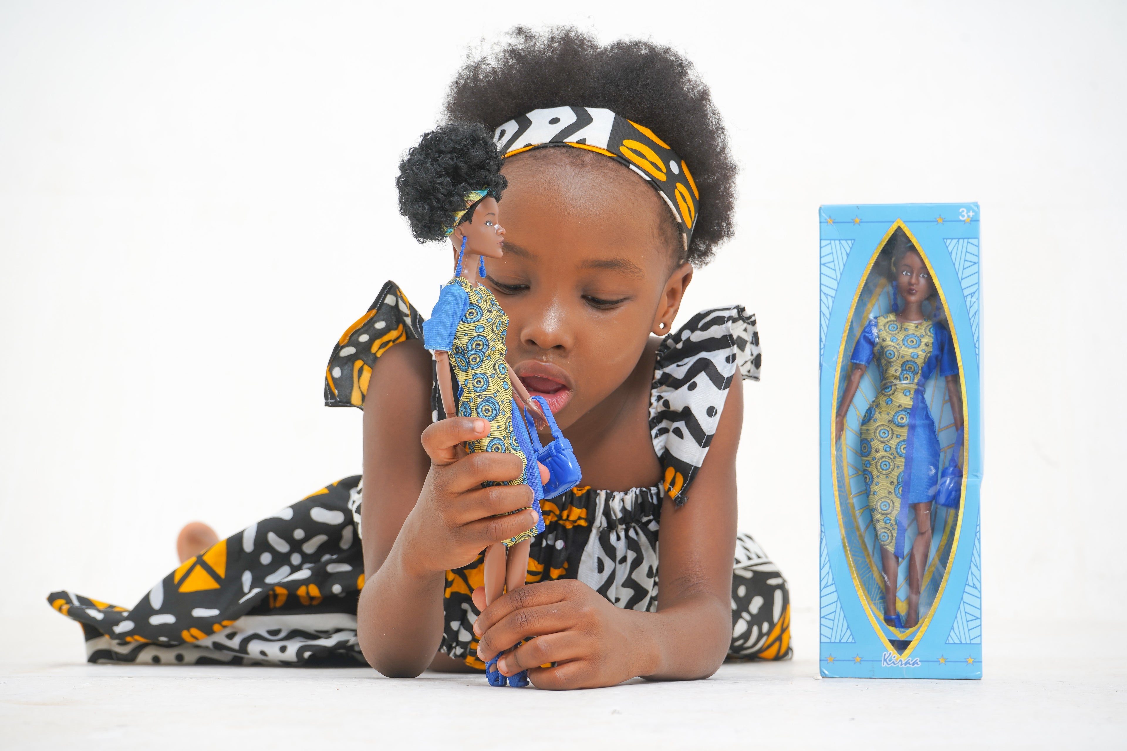 Black girl playing with the Kiraa Black doll with the doll box beside her.